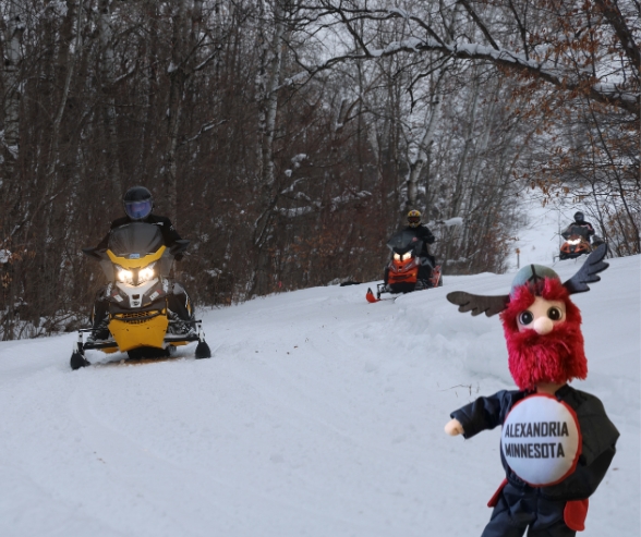 Three people ride snowmobiles along DATA Trails through a snowy forest, while a red and black Little Big Ole mascot doll with "Alexandria Minnesota" on its shirt stands in the foreground.