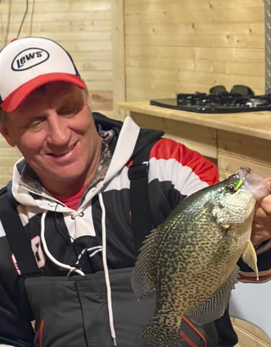 A smiling man in a red and white cap holds up a large crappie fish indoors, wearing a black and red jacket. Light wooden walls and a stove are visible in the background—a perfect moment to share your favorite fishing tip.