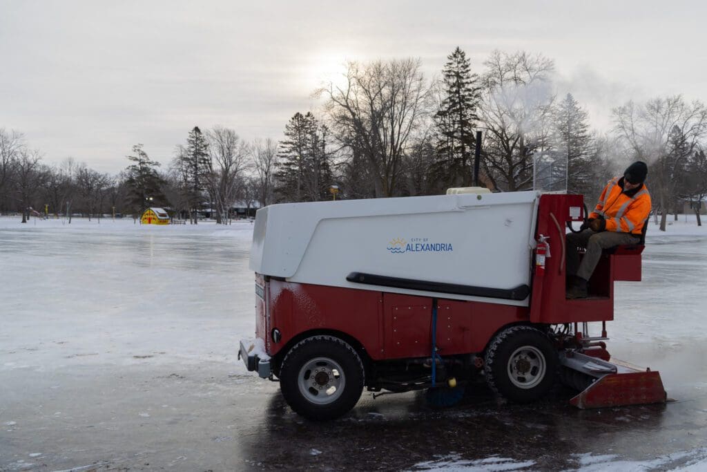 A person in an orange safety jacket drives an ice resurfacing machine across an outdoor skating rink, with bare trees, snow, and a yellow building in the background under a cloudy sky.