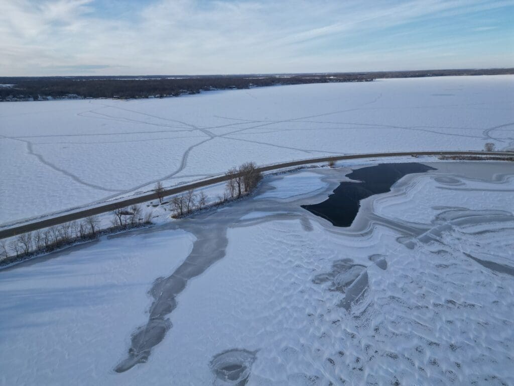 A frozen lake in the Alexandria Lakes Area features snow-covered ice, intersected by a road; winding tire or walking tracks cross the snowy surface, with open water near the shore and a partly cloudy blue sky above.