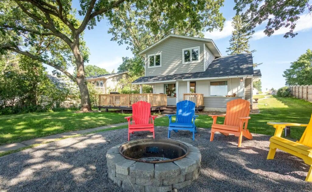 A backyard with a stone fire pit surrounded by colorful Adirondack chairs on gravel, perfect for relaxing after enjoying Lake Henry views. In the background, a two-story house with a large wooden deck is shaded by tall trees on a sunny day.