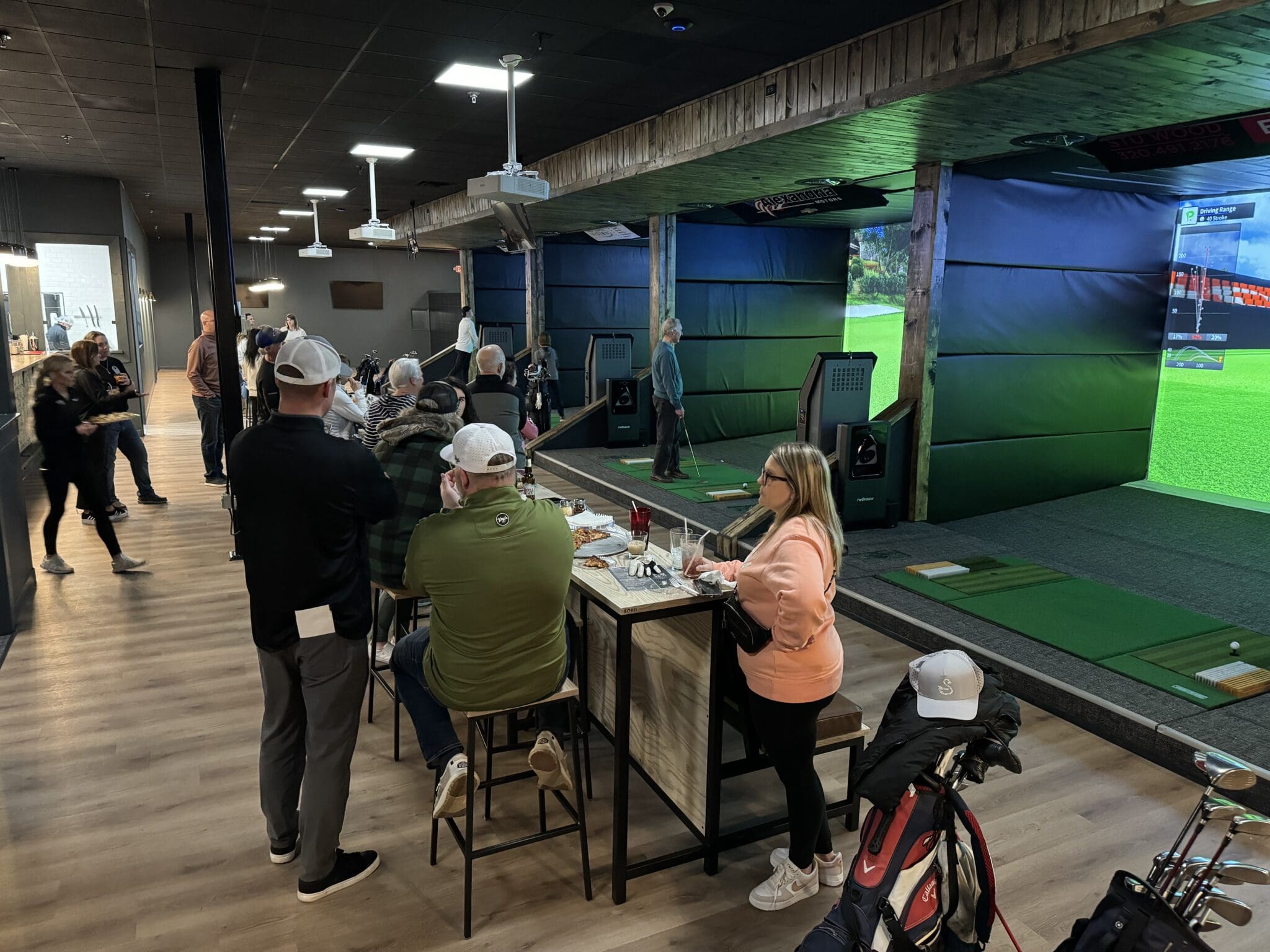 People gather at an indoor golf facility with Hook and Slice simulators, some playing and others seated at tables socializing. Golf bags are visible, and a large screen displays a golf course scene. The atmosphere is casual and lively.