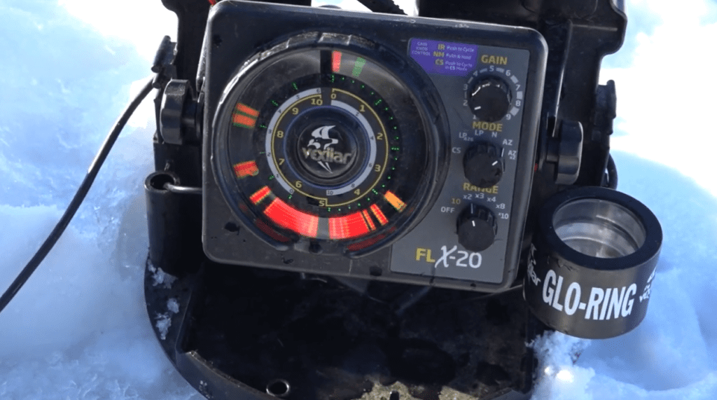 A Vexilar FLX-20 fish finder sits on the snow, displaying colorful sonar readings ideal for winter fishing. Various controls and a Glo-Ring accessory are visible beside the device.