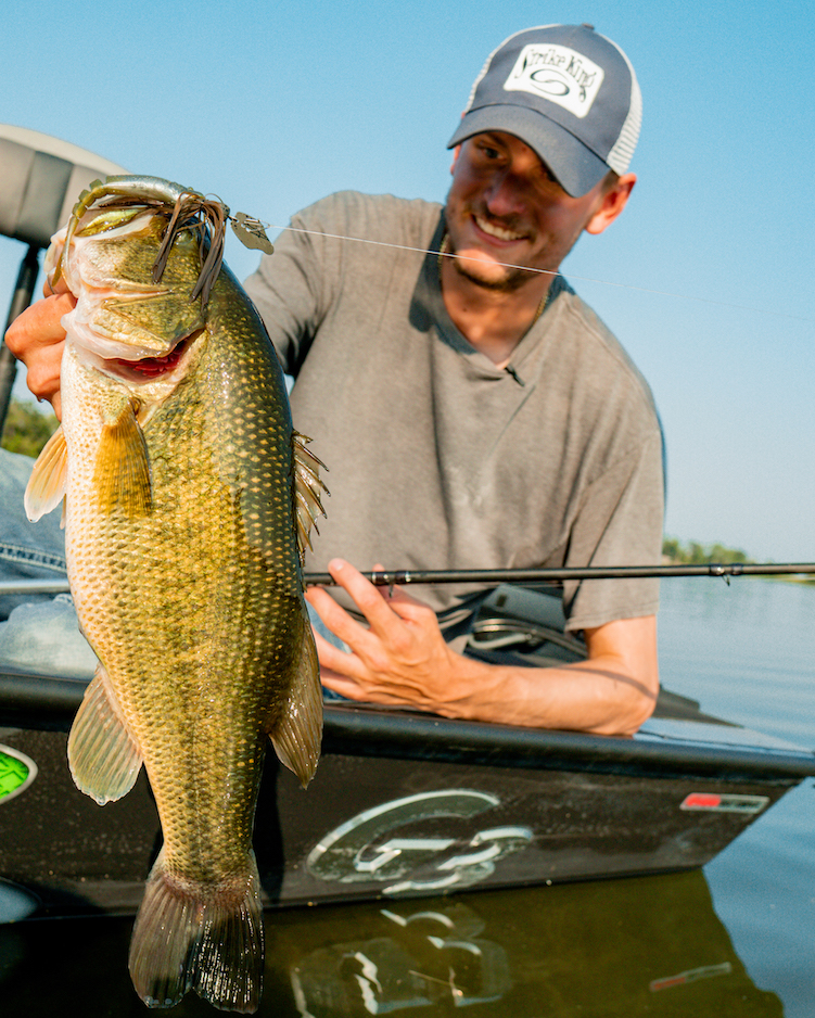 A person in a boat holds up a large bass fish in one hand, smiling at the camera after a fishing success. The person wears a hat and gray shirt, with open water and trees visible in the background.