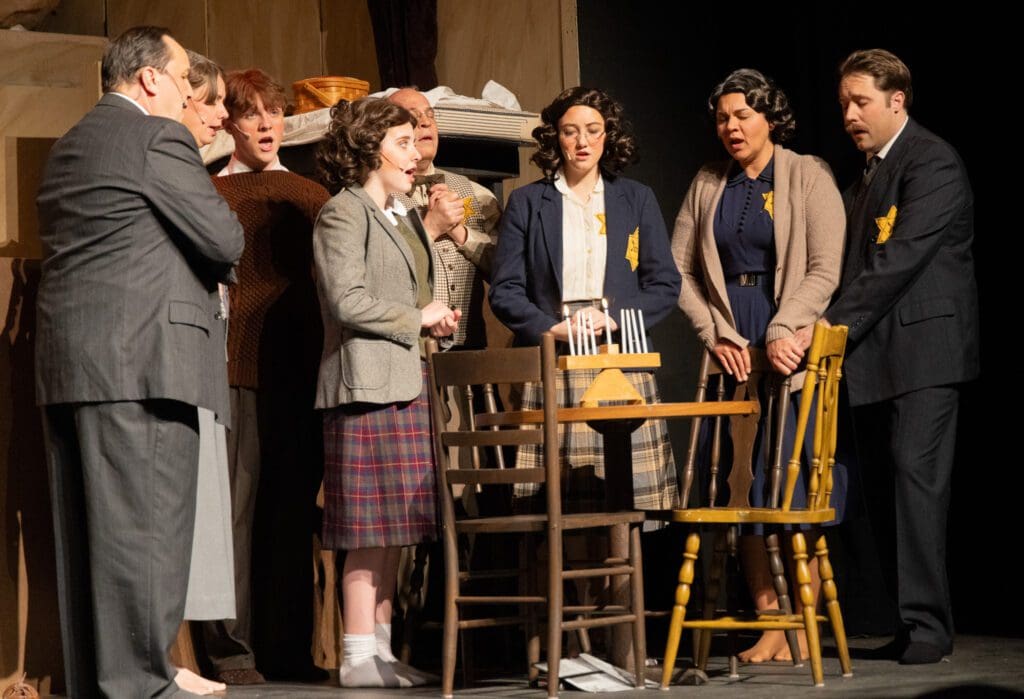 A group of actors in period clothing stands closely together around a table on the Andria Stage, appearing serious and contemplative—some wear yellow Star of David patches, evoking a powerful scene from The Diary of Anne Frank.