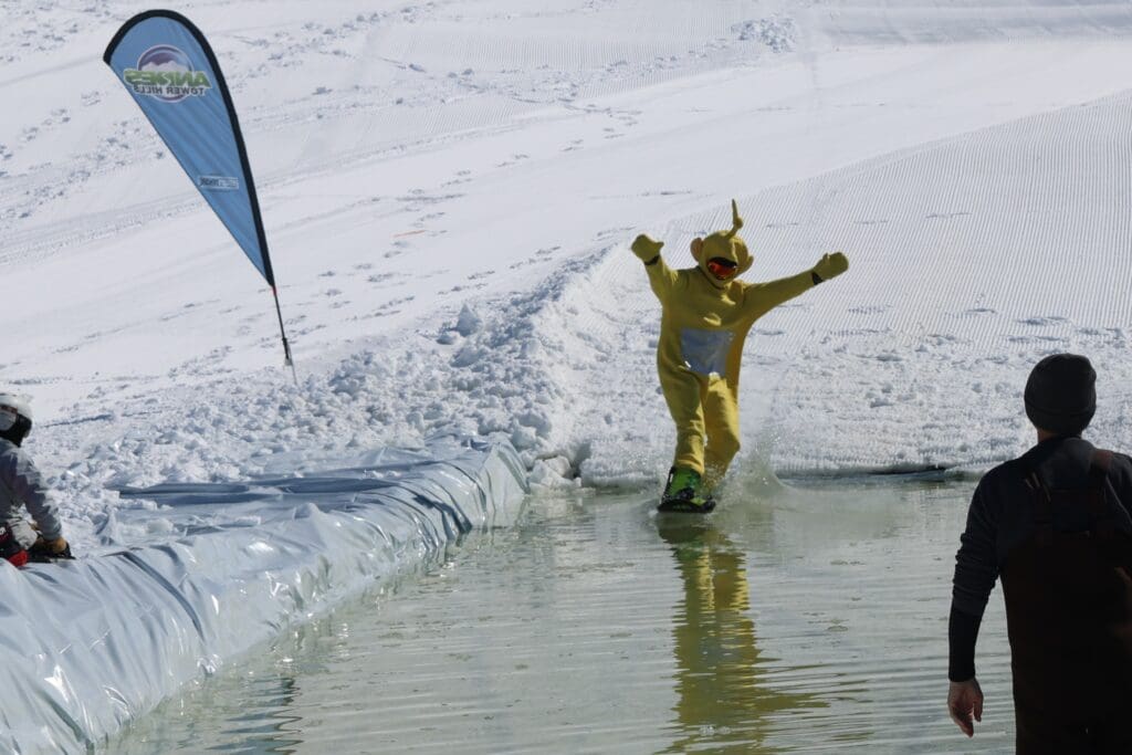 A person in a yellow Teletubby costume snowboards across a pool of water on a snowy slope at Andes Tower Hills during the End of the Season Bash, while a few spectators watch nearby.