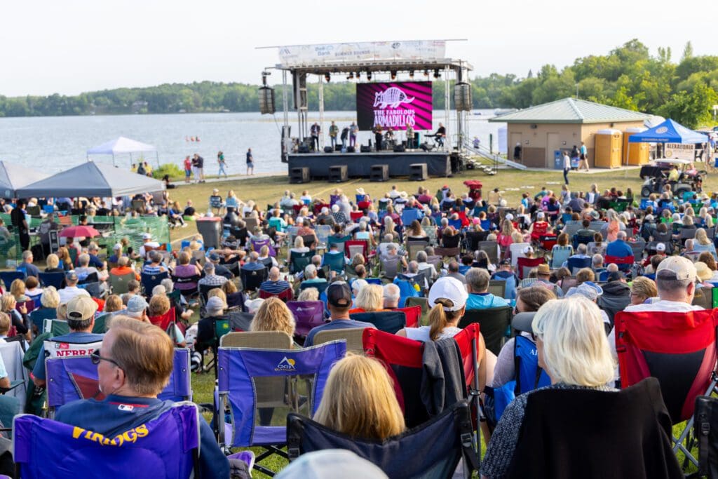A large crowd sits in lawn chairs facing an outdoor stage by a lake, watching a live Summer Sounds performance. Trees and a small building are in the background, with a large screen on the stage under sunny, clear weather.