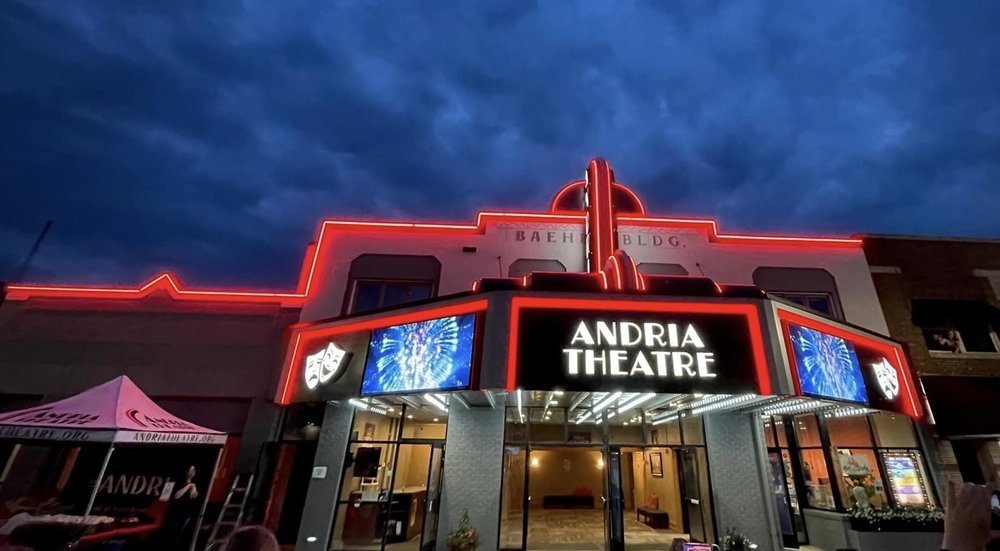 The Andria Theatre building at night, outlined with red neon lights, glows under a dramatic dark blue sky. The Andria Theatre’s entrance is lit up, with digital screens displaying graphics on both sides of the theater sign.