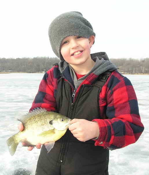 A smiling child in a gray beanie and red plaid jacket holds a fish while standing on a frozen lake, with bare trees in the background—another memorable catch with JS Guide Services.