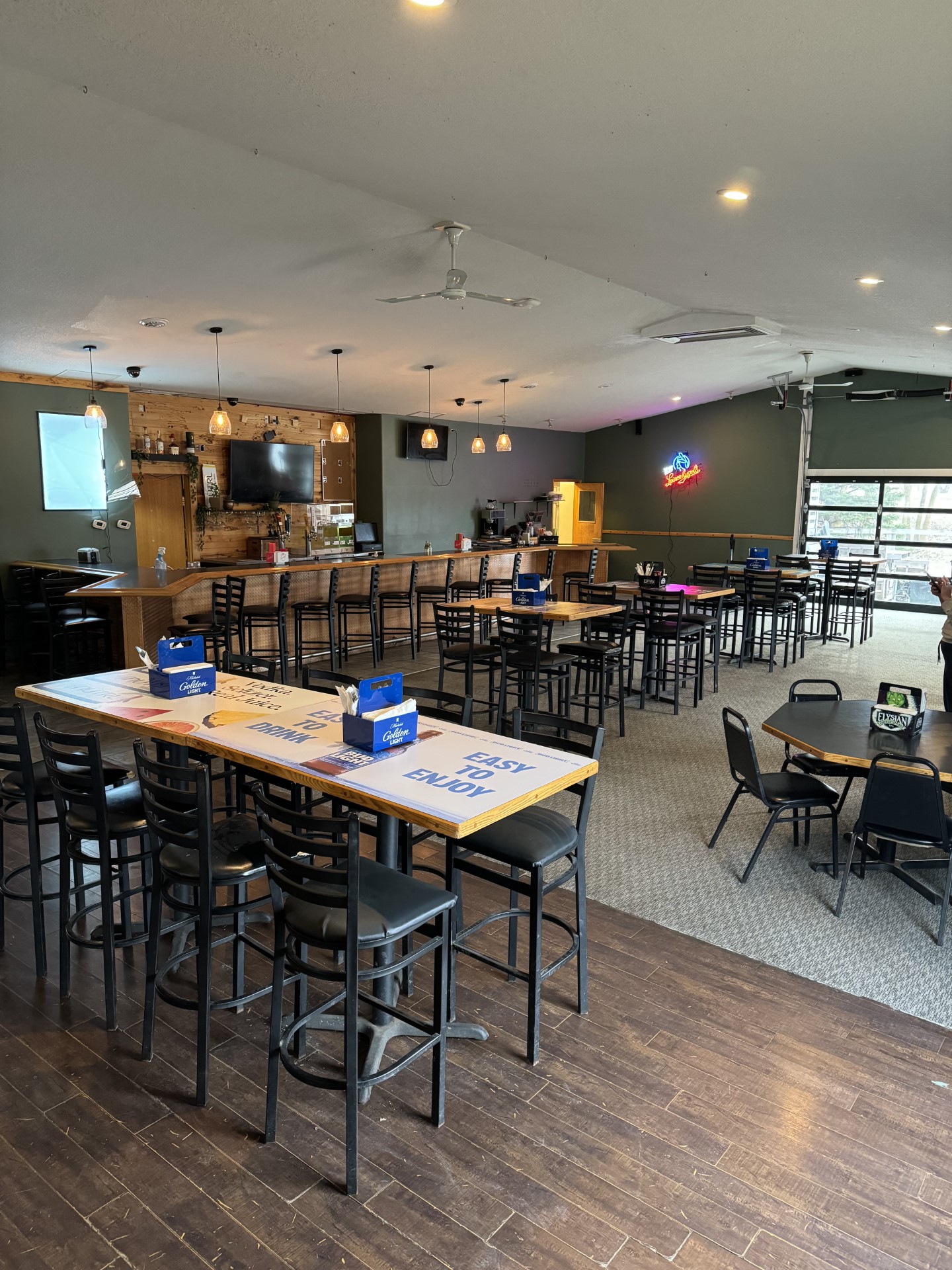 Spacious bar and restaurant interior with high tables and chairs, a long bar counter, TV, neon signs, and large windows providing natural light. The Locals offers clean, modern Dining in one of the top Alexandria restaurants, warmly lit for a welcoming vibe.
