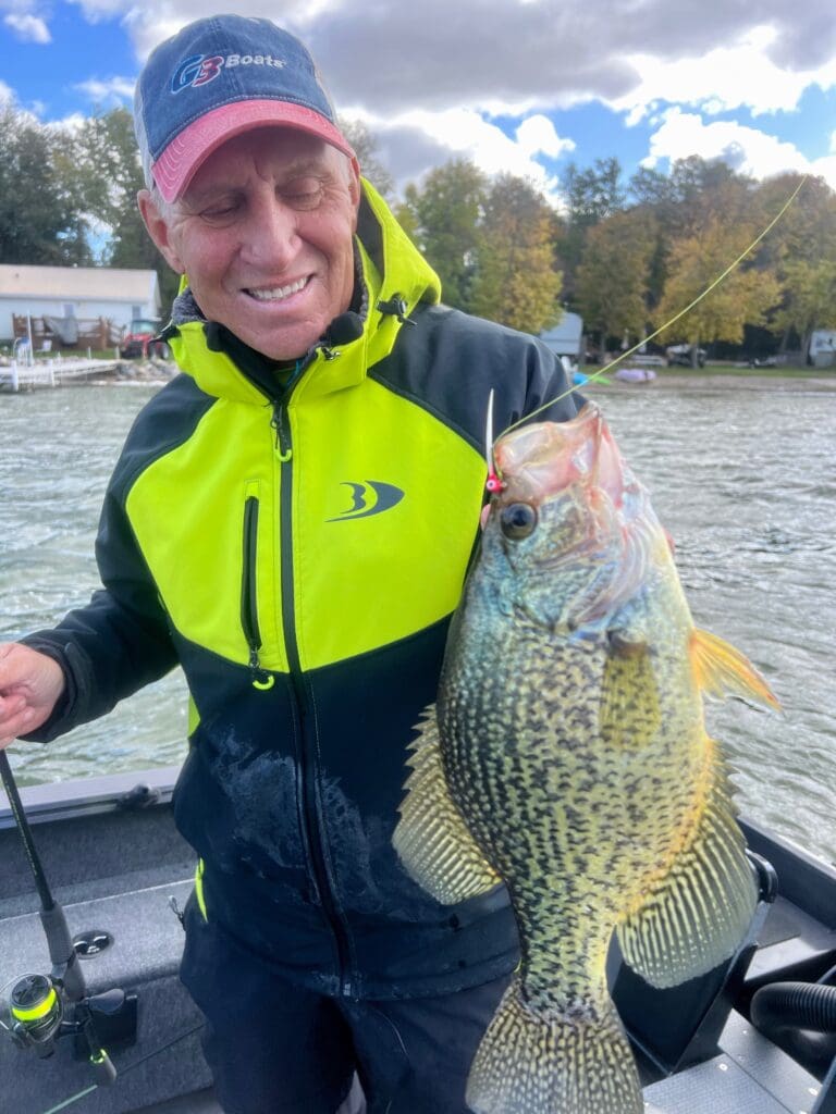 A smiling man in a yellow and black jacket holds up a large crappie fish on a fishing line while fishing the Midwest from his boat on a lake, with trees and houses visible in the background.