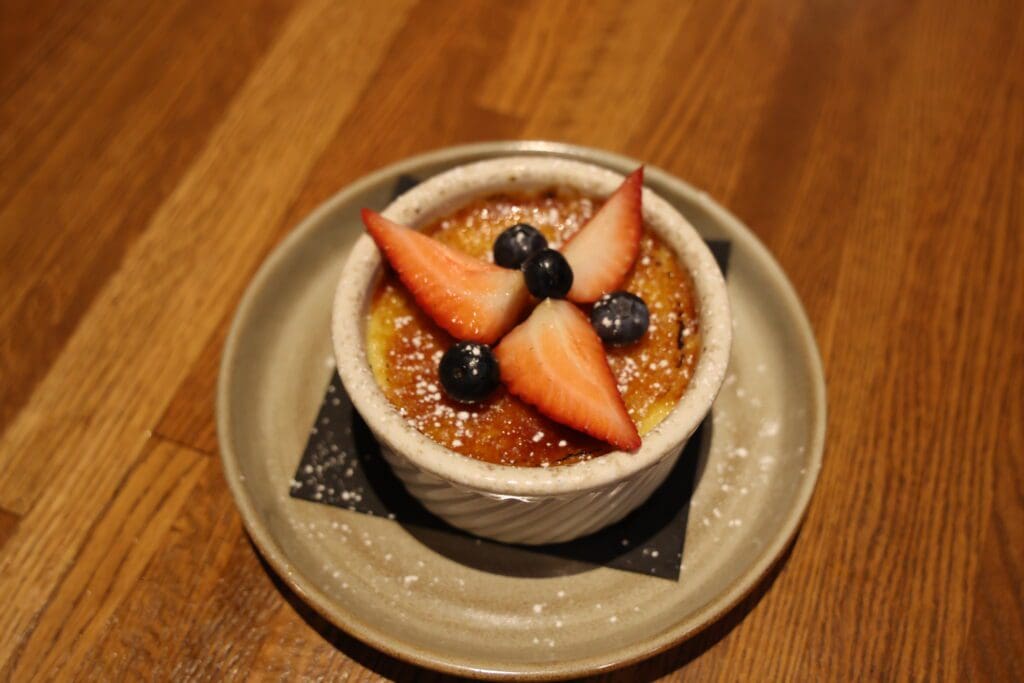 A ramekin of crème brûlée topped with strawberry slices and blueberries, lightly dusted with powdered sugar, sits on a beige plate—an elegant dessert often found at top MN Restaurants in Alexandria.