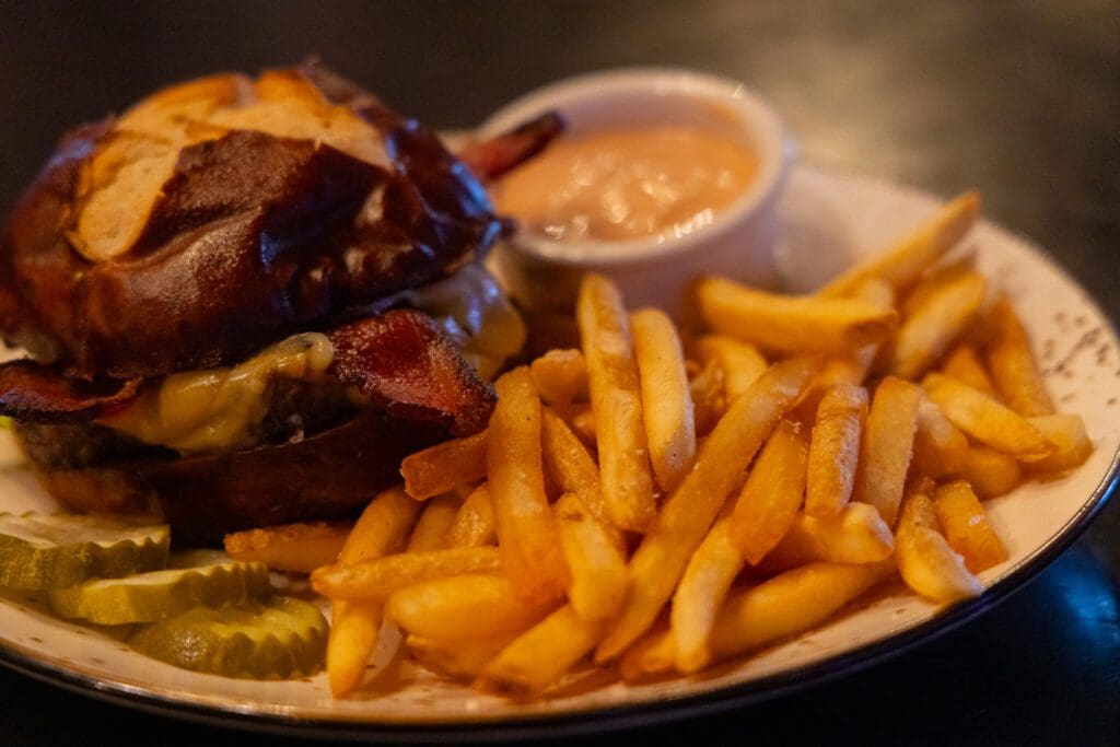 A plate with a cheeseburger on a pretzel bun, crispy French fries, pickle slices, and a small cup of dipping sauce—classic comfort food you’ll find at top Alexandria MN restaurants.