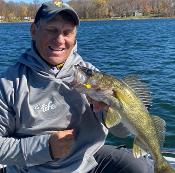 A smiling person in a gray hoodie and baseball cap holds up a large yellow-green fish on a boat, enjoying fishing the Midwest on one of Alexandria MN’s picturesque lakes, with autumn foliage and a blue lake in the background.