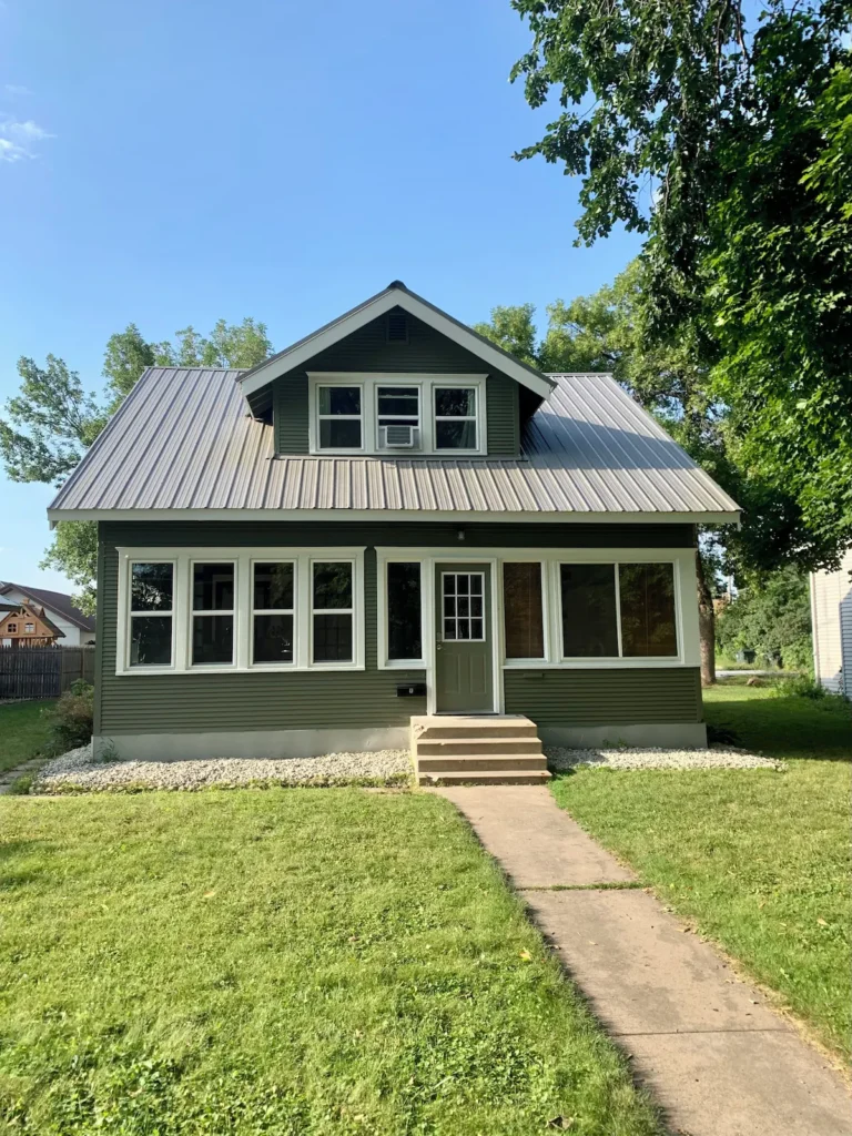 The Boho Bungalow is a green two-story house with a gray metal roof, large front windows, and a small front porch with steps, set on a grassy lawn with trees and a sidewalk leading to the door.