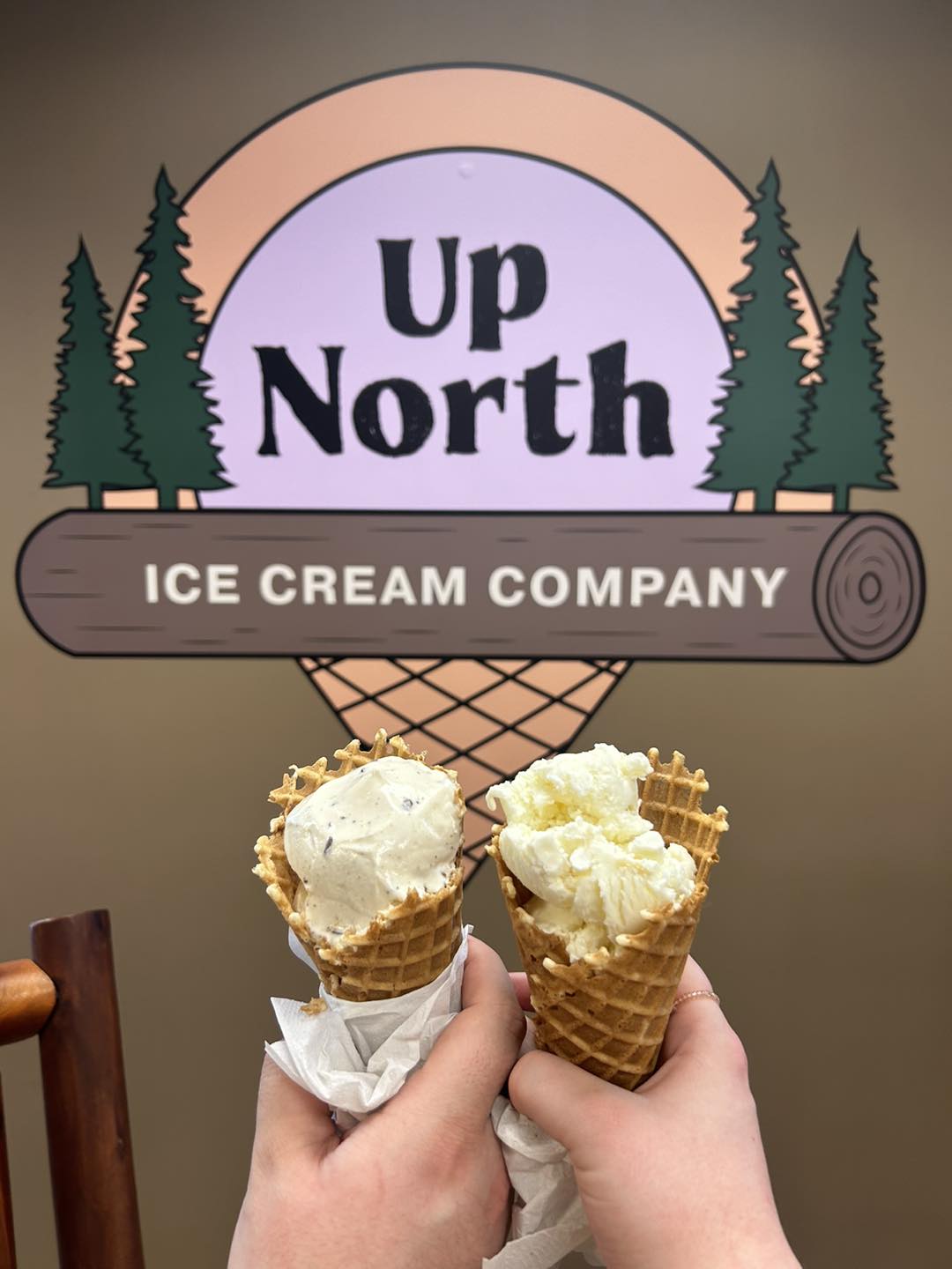 Two hands hold waffle cones topped with creamy scoops, lifted in front of the Up North Ice Cream Company sign, which features a rustic log and pine tree design.