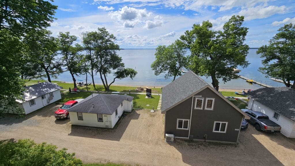 View of lakeside cabins and parked cars with trees and people on a grassy area by the beach at Viking Bay Resort; calm lake water and a dock stretch into the distance under a partly cloudy sky.