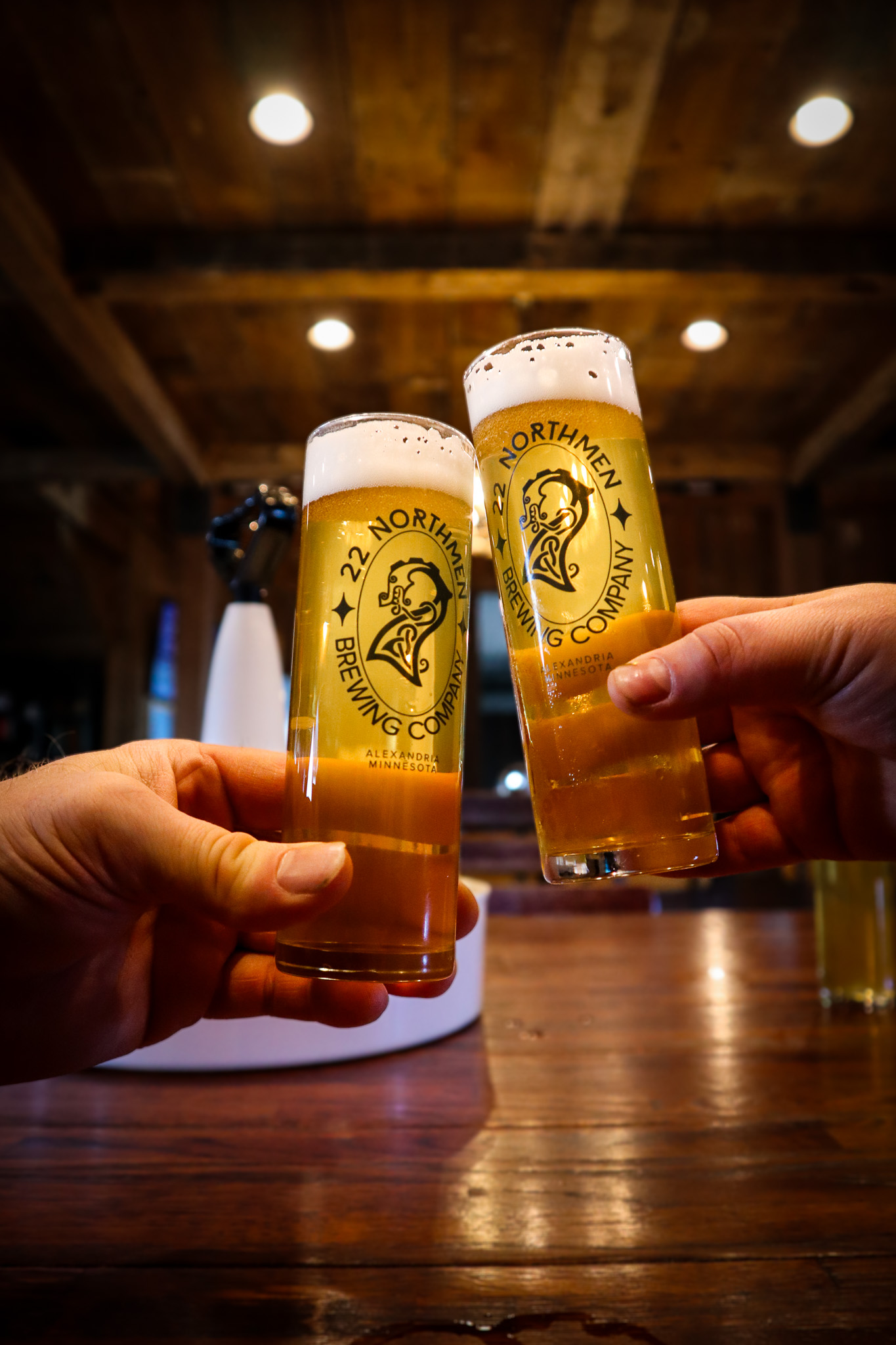 Two hands clink tall beer glasses with "22 Northmen Brewing Company" logos, stoking the spirit of the brewery against a rustic wooden backdrop. Both glasses are filled with golden beer and crowned with a frothy head.