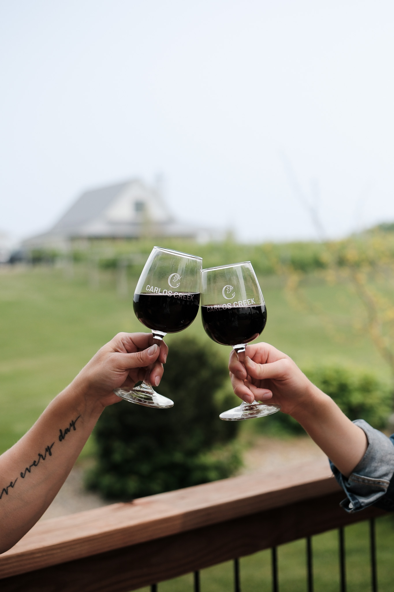 Two people clink glasses of red wine on a wooden deck at Carlos Creek Winery, with a blurred view of a vineyard and house in the background. One person has a tattoo on their forearm.