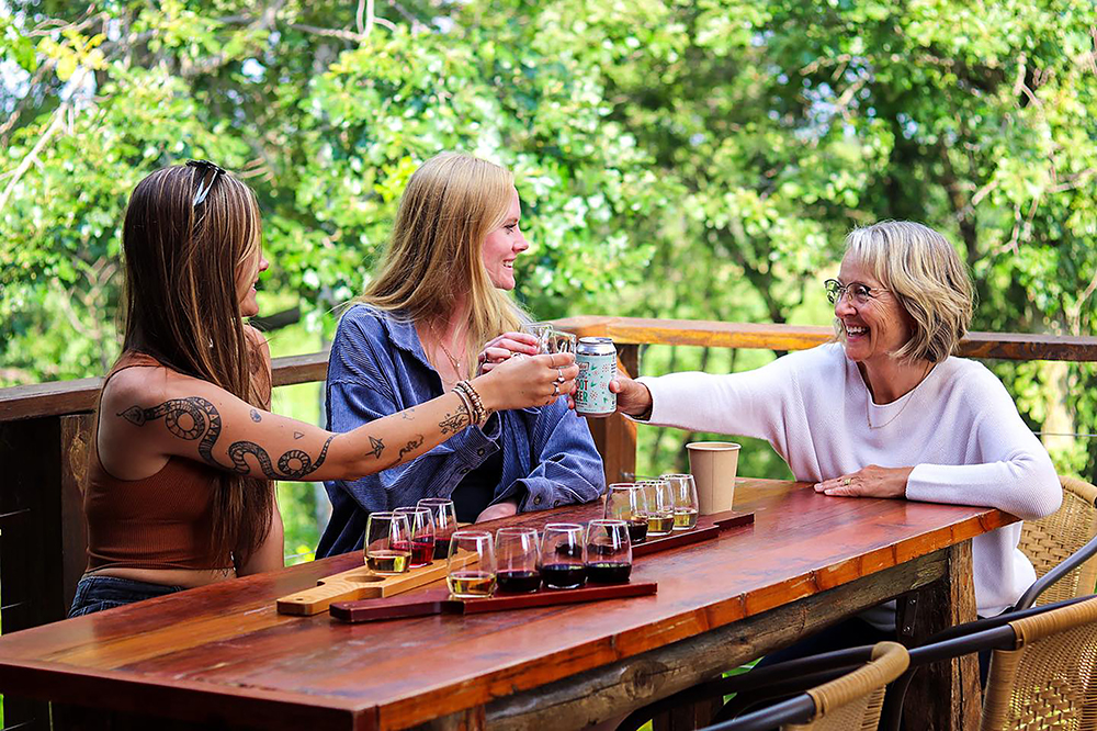 Three women sit at an outdoor wooden table at Burr Vineyards, smiling and toasting drinks together, with flight boards of beverages in front of them and lush green trees in the background.