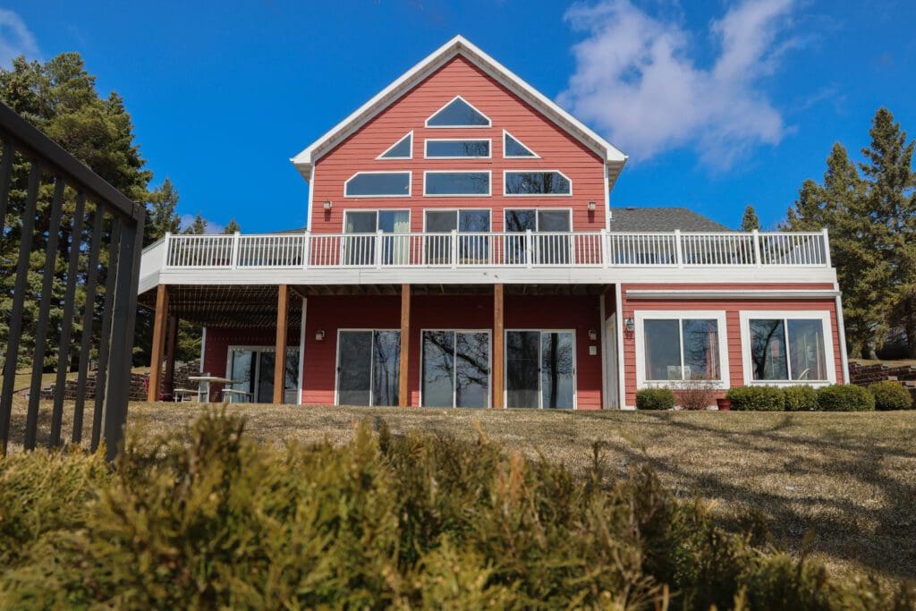 A two-story red chateau with white trim and large windows, featuring a spacious upper deck and a grassy lawn, surrounded by trees under a bright blue sky with scattered clouds.