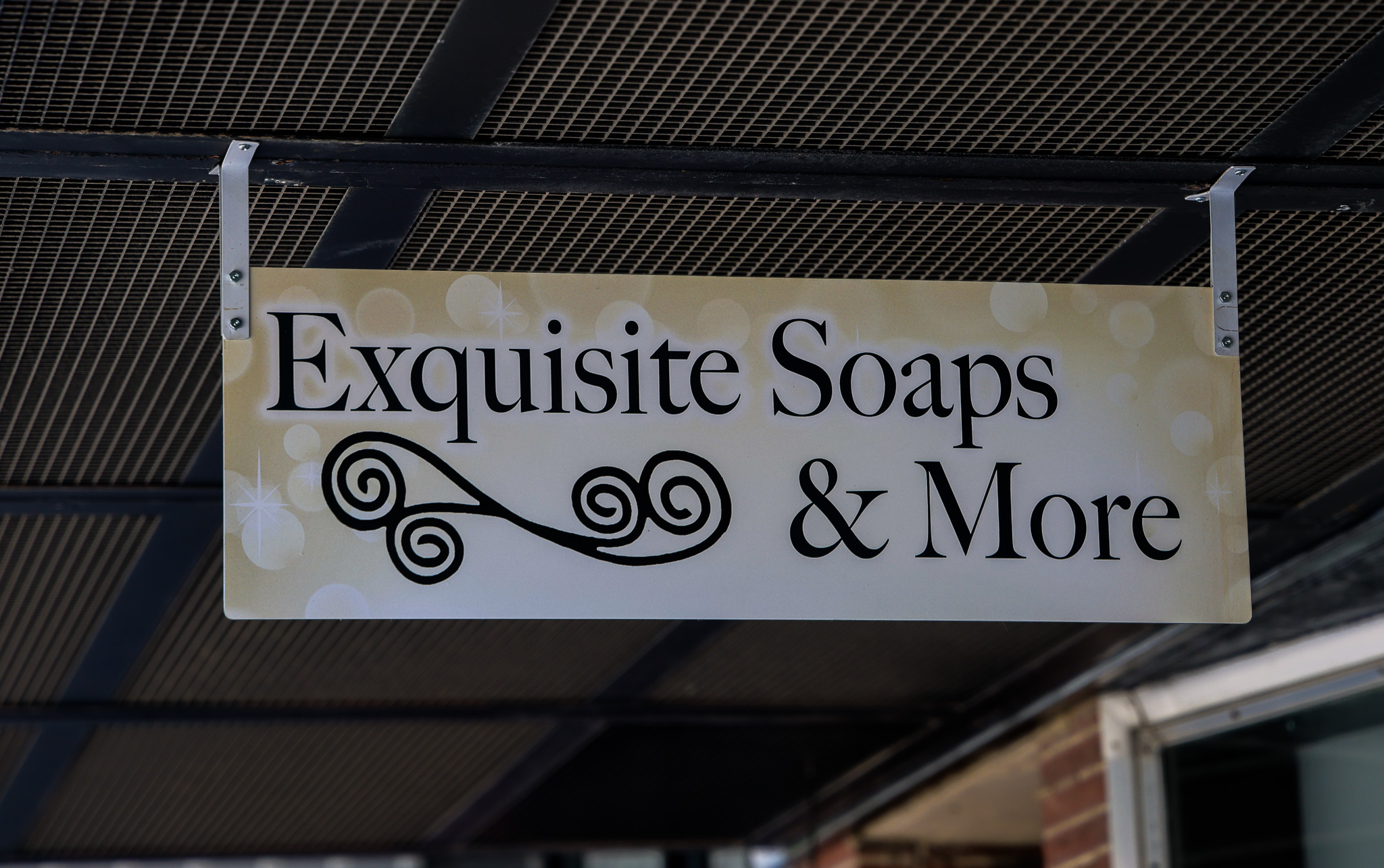 A rectangular shop sign reads "Exquisite Soaps & More" in bold black text with decorative swirls, perfectly capturing the charm of Exquisite Soaps, and hangs elegantly under a metal awning.
