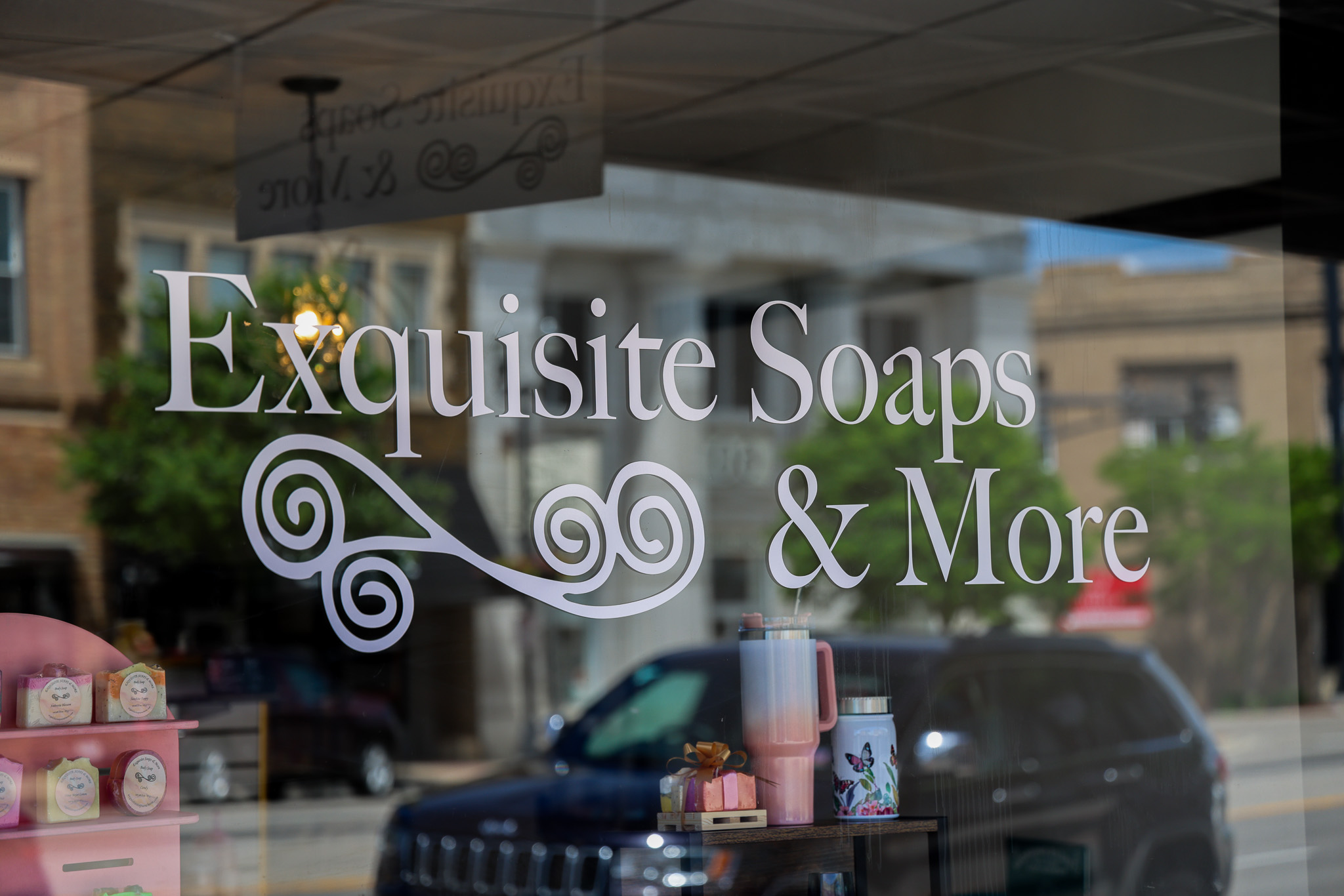 Storefront window with "Exquisite Soaps & More" in white lettering and a decorative swirl design, reflecting the street, nearby buildings, and a parked car—perfectly capturing the charm of Exquisite Soaps.