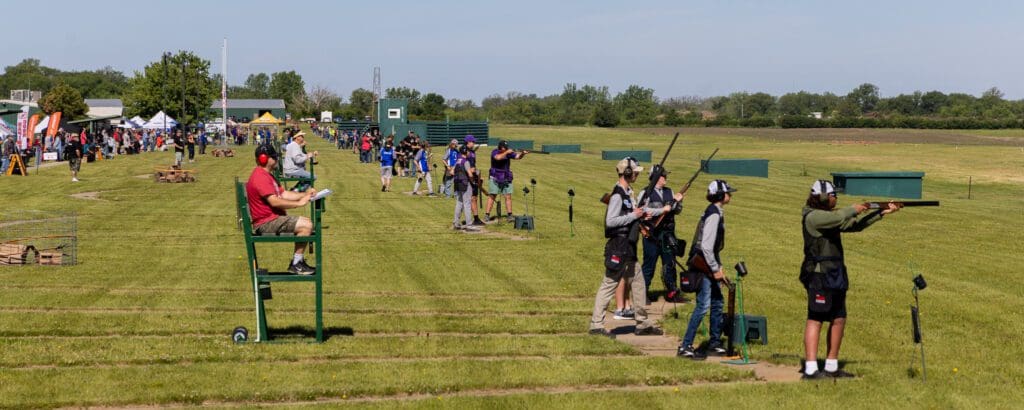 Student athletes participate in the Alexandria Trap Shooting Championship at an outdoor range. Shooters, judges, and spectators line several lanes on a grassy field under sunny skies with trees in the background.
