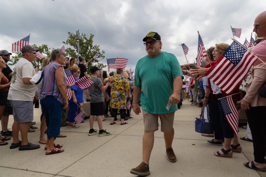 A man in a green shirt and veteran hat walks past a crowd holding American flags outdoors on a cloudy day in Alexandria. People line the sidewalk, reaching out and watching as the Vietnam Veteran receives a heartfelt Welcome Home.