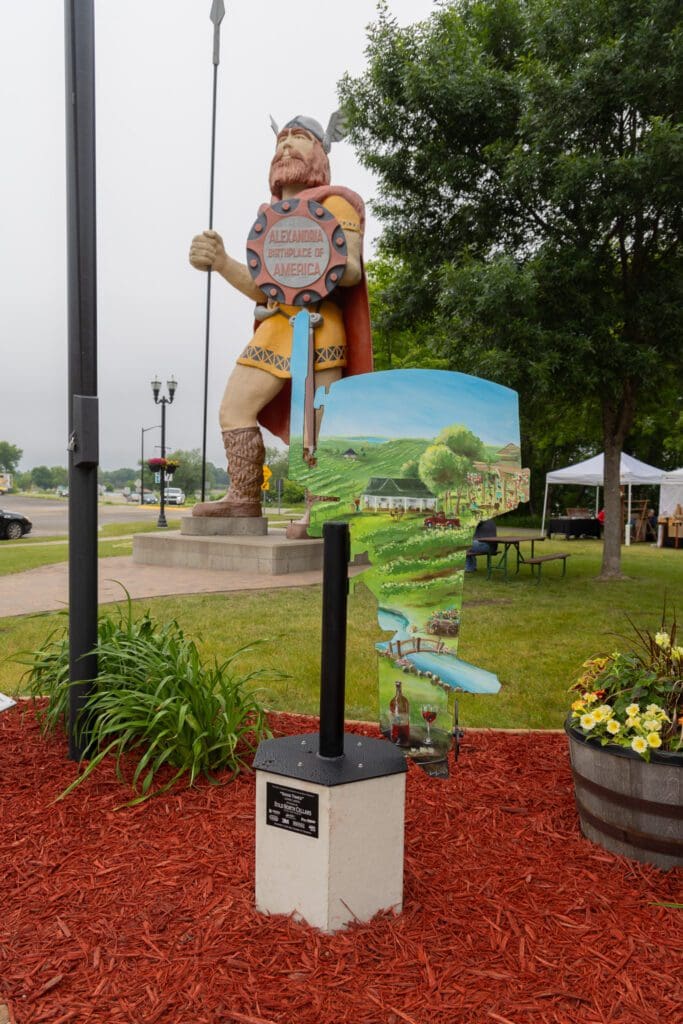 A large Viking statue stands behind a painted Minnesota-shaped sign on red mulch, surrounded by plants and flowers, in a park with green grass and trees—find it easily with our interactive map.