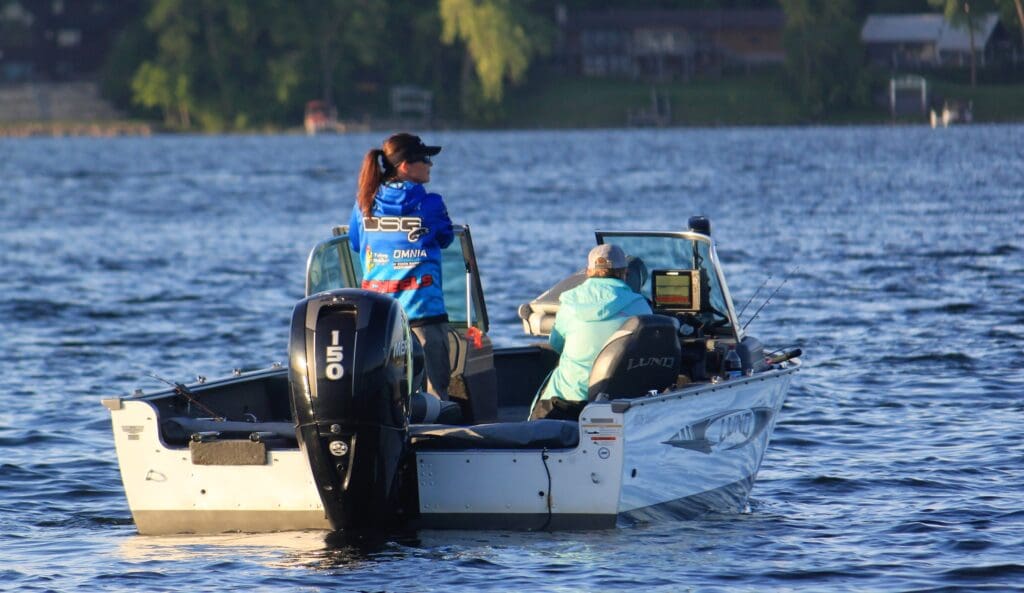 Two women anglers are fishing on a small white motorboat on a Minnesota lake. One stands near the motor while the other sits facing a screen. Trees and buildings line the distant shoreline, evoking an open water tournament atmosphere.