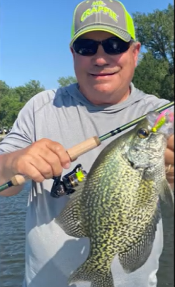 A smiling man in sunglasses and a bright cap holds a fishing rod and proudly displays a large crappie he caught while fishing, standing by the water with trees in the background.