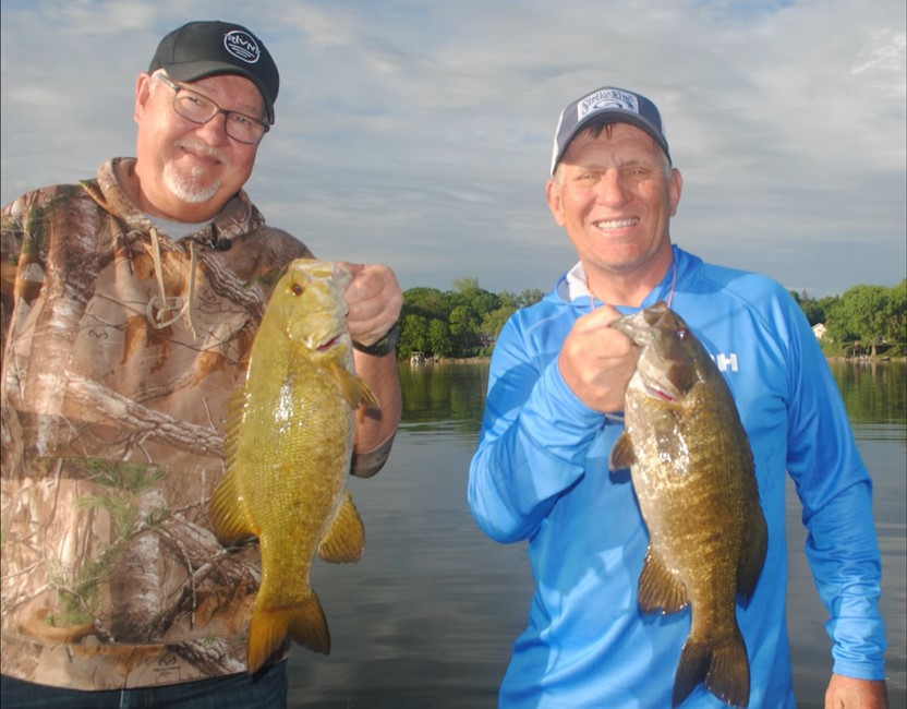 Two smiling men in outdoor clothing and hats proudly hold up large fish while standing near a calm lake—showing off how to catch more summer bass with ease amid trees and houses in the background.