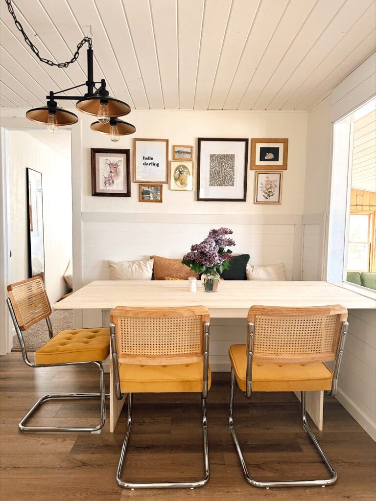 The Darling Cabin’s dining area features a light wooden table, three yellow-cushioned chairs, wall art above, a vase of purple flowers, and pendant lights on a white paneled ceiling. Natural light pours in from the window.