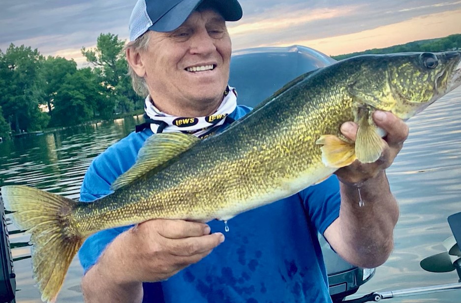A smiling man in a blue shirt and cap proudly holds a large, freshly caught walleye in both hands while standing on a boat on a calm lake, with trees and a cloudy sky in the background.