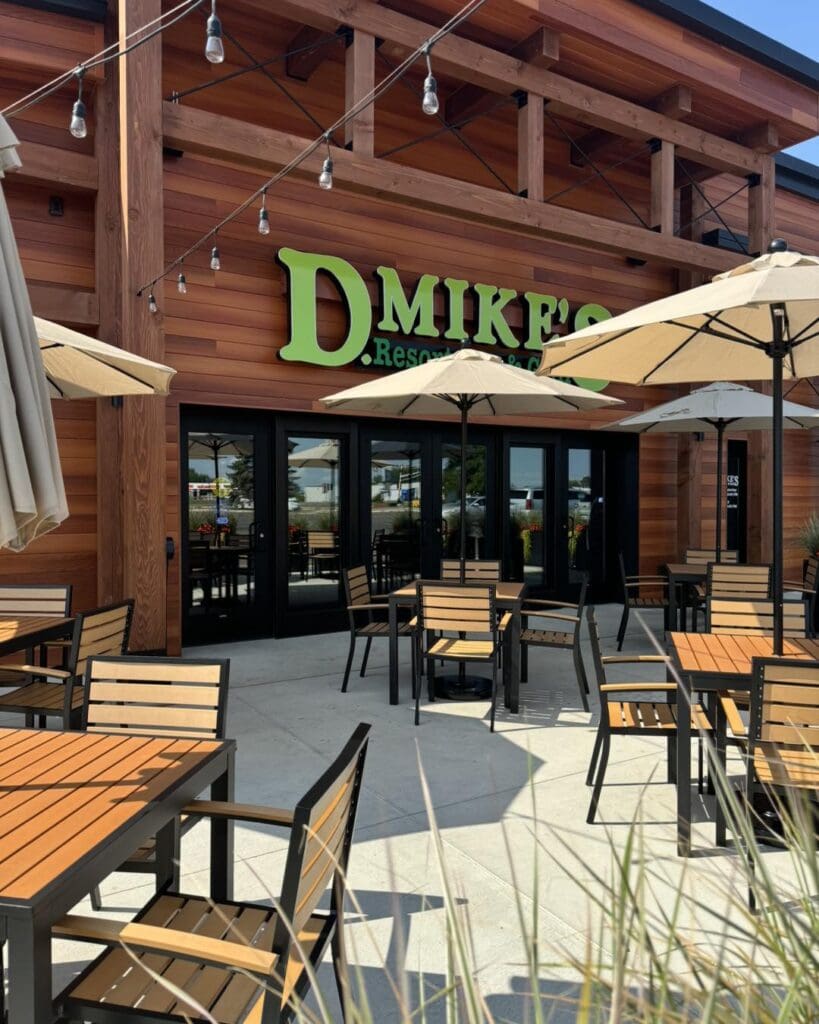Outdoor patio of "D. Mike's" Grill with wooden tables and chairs under beige umbrellas; string lights hang above and the building has wood paneling and large glass doors, giving a relaxed Resort Bar vibe.