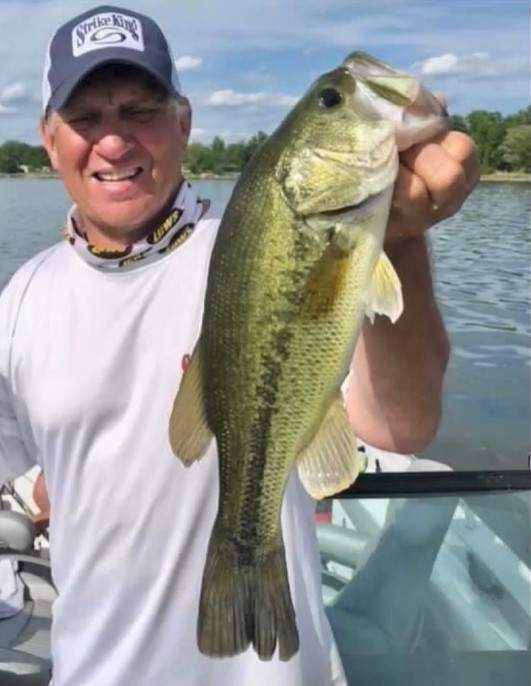 A man wearing a cap and white shirt smiles while holding a large bass fish in one hand, standing on a boat with water and trees in the background—enjoying fishing weed lines during the dog days of summer.