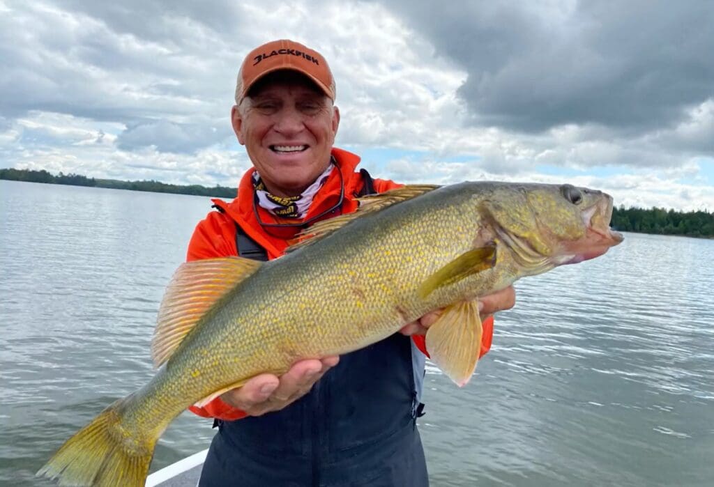 Smiling man in an orange cap and jacket, possibly Mike Frisch, holds a large walleye with yellowish scales while standing on a boat in a lake, with trees and cloudy sky in the background.