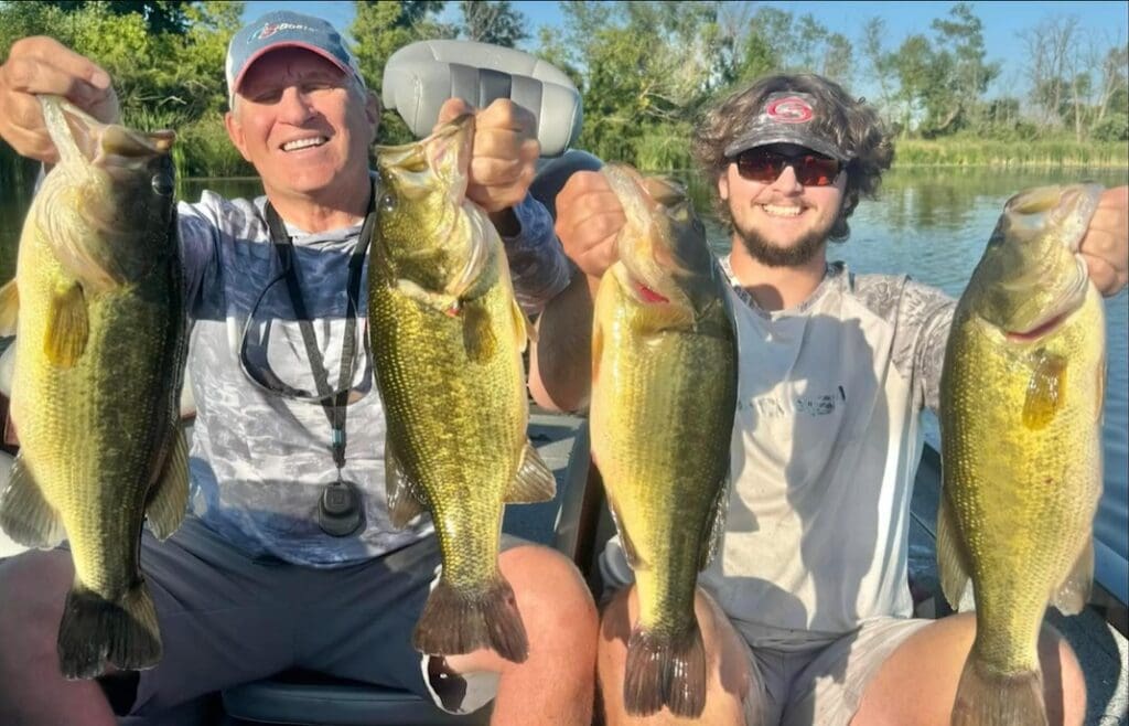 Two people sitting in a boat on a lake, smiling and proudly holding up four large fish they caught after an exciting bass bite, with green trees and blue sky in the background.