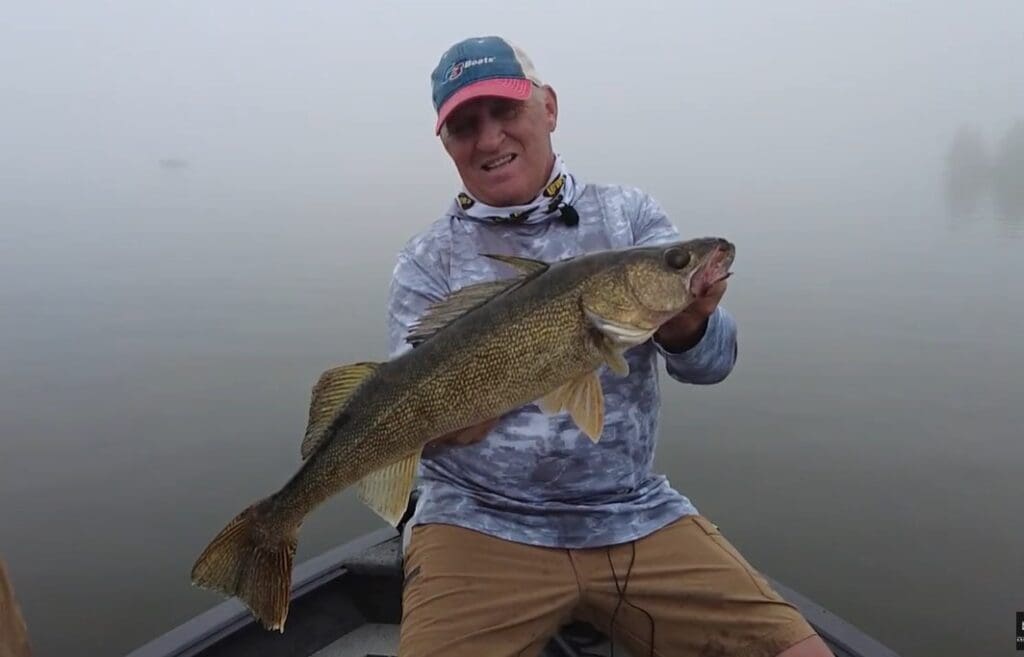 A smiling man in a cap and fishing attire sits in a boat on a foggy lake, proudly holding a large fish with both hands after catching walleyes as the weather cools down.