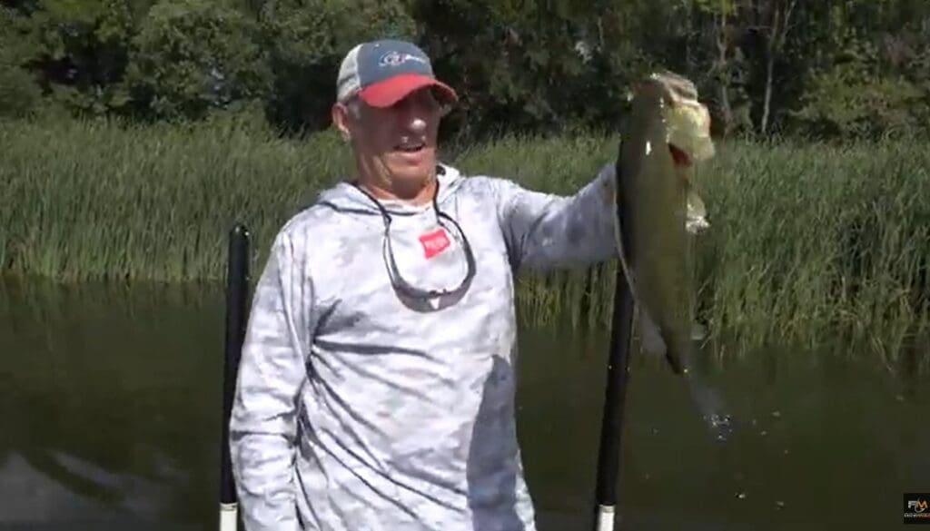 A man in a gray hoodie and red cap holds up a large fish next to a body of water in the Alexandria Lakes Area, with tall grass and trees in the background.