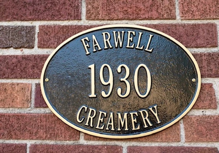 An oval metal plaque mounted on a brick wall reads "FARWELL CREAMERY 1930" in capital letters, with a gold border around the edge, marking the legacy of The Creamery & Corner Garage.