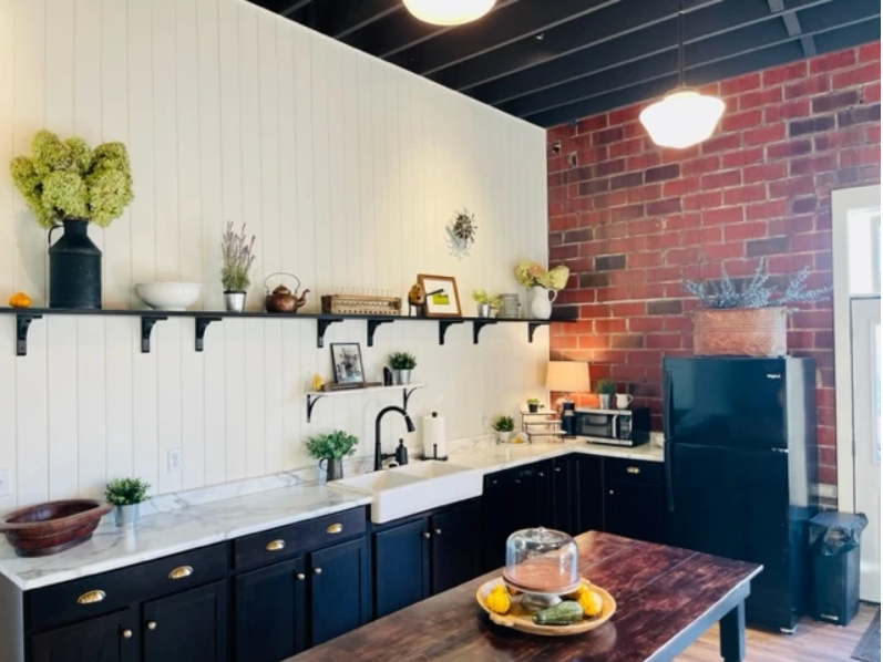 A modern kitchen at The Creamery & Corner Garage features dark cabinets, white countertops, exposed brick walls, open shelves with plants and kitchenware, black appliances, and a wooden table with a cake stand and lemons.