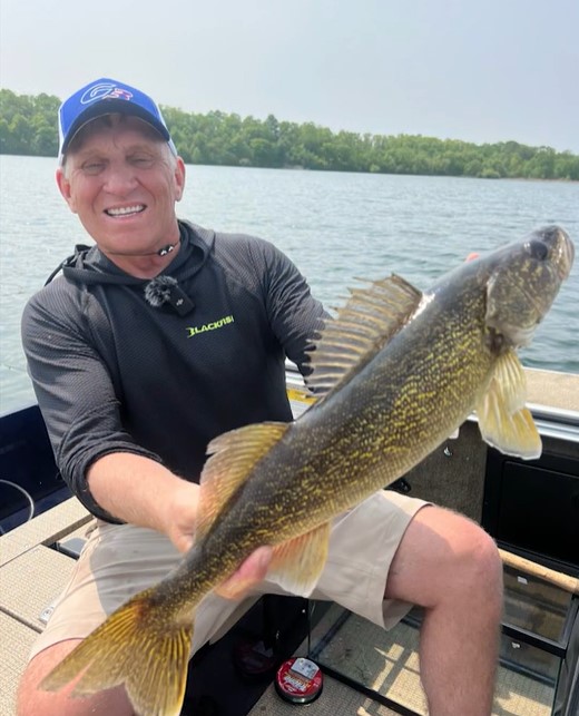 A smiling man in a cap and black shirt sits on a boat, proudly holding a large fish with yellow and brown markings—caught using Multi-Species Swimbaits—with a scenic lake and trees in the background.