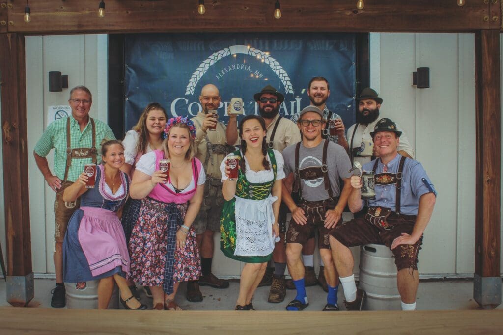 A group of people dressed in traditional Bavarian clothing smile and hold beer glasses, posing together in front of an Oletoberfest banner at a festive event.