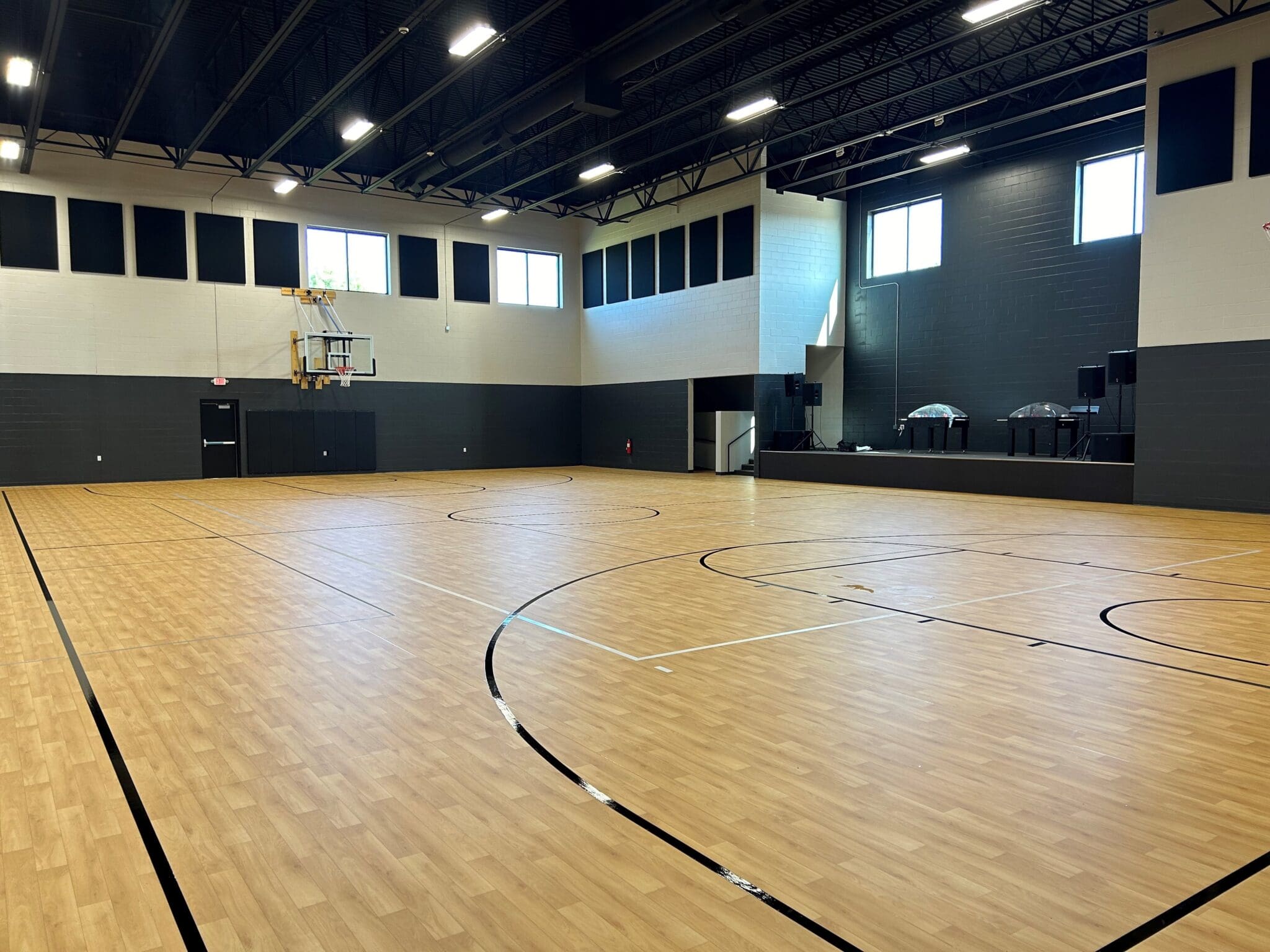 An empty indoor basketball court with a wooden floor, black lines, and high windows. There is a basketball hoop at one end, a stage area with speakers and tables on the right, and room for pickleball games—part of the versatile VIE Church gym.