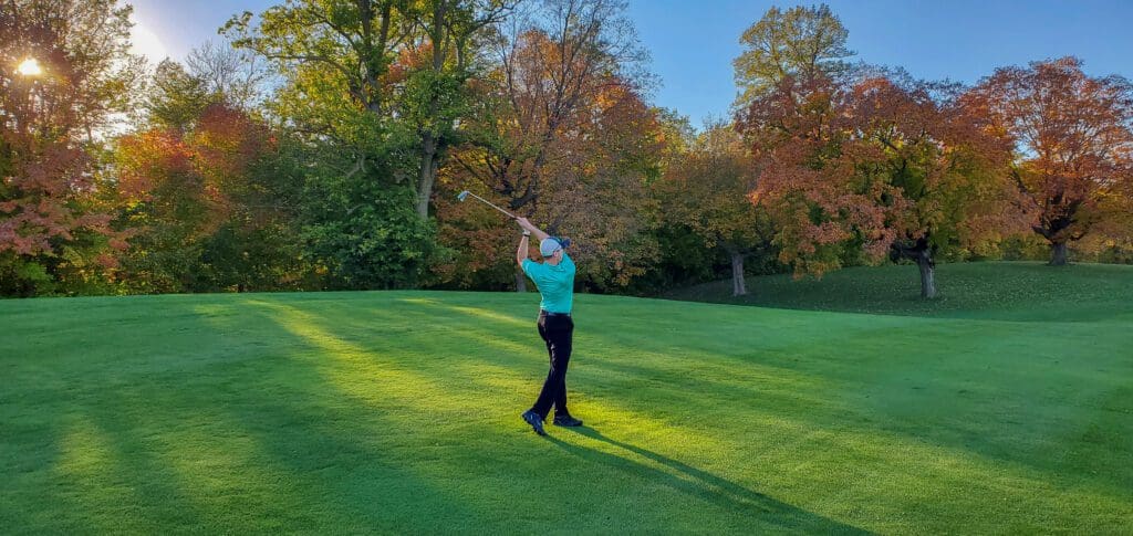 A golfer swings a club on a green course surrounded by colorful autumn trees in the late afternoon sunlight, savoring the peaceful scene as some golf courses are closing down.