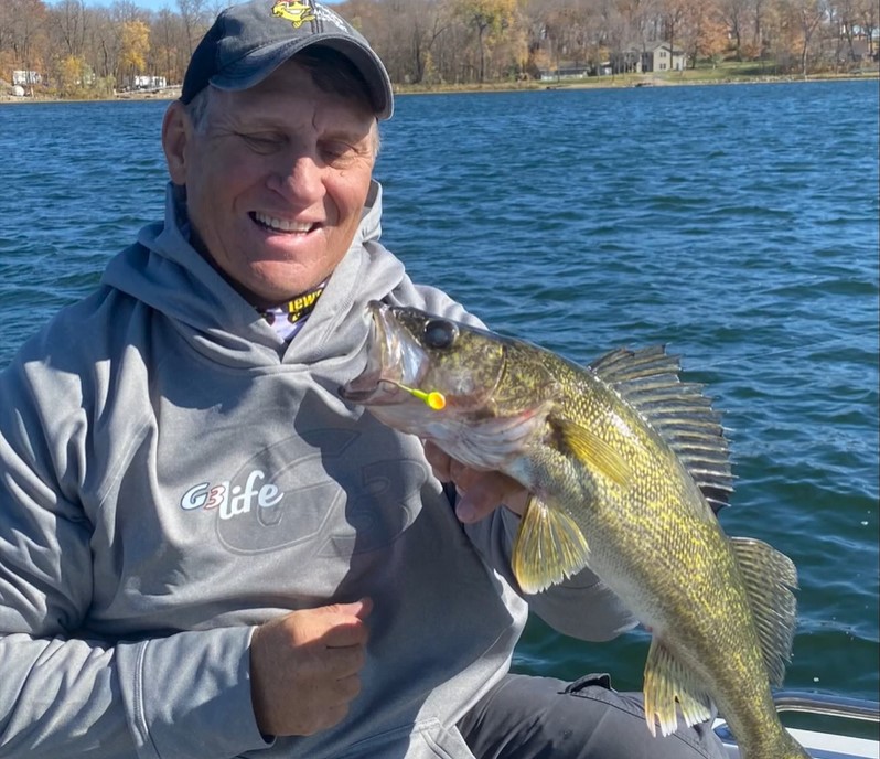 A smiling person in a gray hoodie and cap holds a large yellow-green fish with a lure in its mouth while enjoying perfect fall fishing weather from a boat on a blue lake. Trees with autumn leaves are visible in the background.