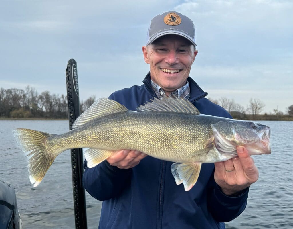 A smiling man in a blue jacket and cap holds a large fish in both hands while standing on a boat, capturing the excitement of end of the season fishing with a lake and bare trees in the background.