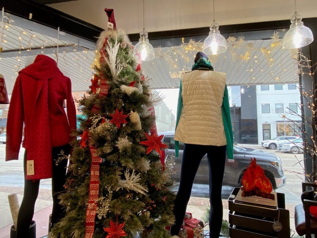 A store window display features two mannequins in winter clothes beside a decorated Christmas tree. Shopping local, you’ll find cozy scenes like this, with a red coat, white vest, faux fireplace, and holiday lights completing the festive atmosphere.